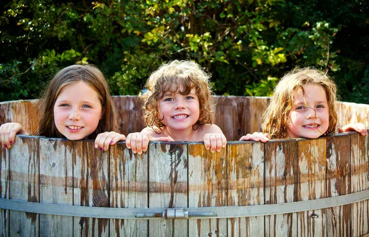 Three girls peeping over a wooden hot tub outside a farm glamping lodge at Hollings Hill